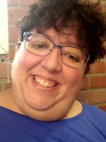Picture of a biracial woman with curly black hair, wearing glasses and a blue blouse in front of a brown brick wall.