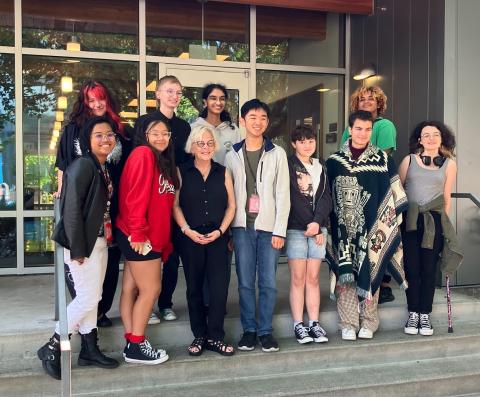 10 LGBTQ Youth council members pose in front of a school building on the stairs with Senator Claire Wilson. 