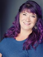 Lisa Keating with dark purple hair, wearing a medium blue crew neck t-shirt, stands smiling in front of a gray vignette background.