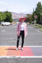 A photo of Desi standing in a rainbow crosswalk wearing a pink jacket and pink cowboy hat. 