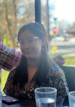 Izzy is sitting a table smiling at the camera. She has long brown hair and glasses. 