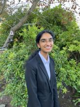 Jaanvi is smiling at the camera in front of green bushes. They have their hair pulled back into a ponytail and are wearing glasses. 