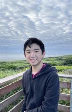 James is smiling at the camera in front of a large field. He is wearing a black jacket. 