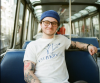 A picture of CJ sitting on a bus, wearing a white t-shirt and blue beanie. CJ wears glasses and is smiling. 